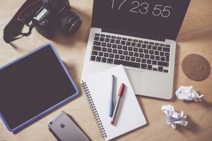 Various items jumbled upon a desktop: notepad, pens, camera, laptop, and iphone.